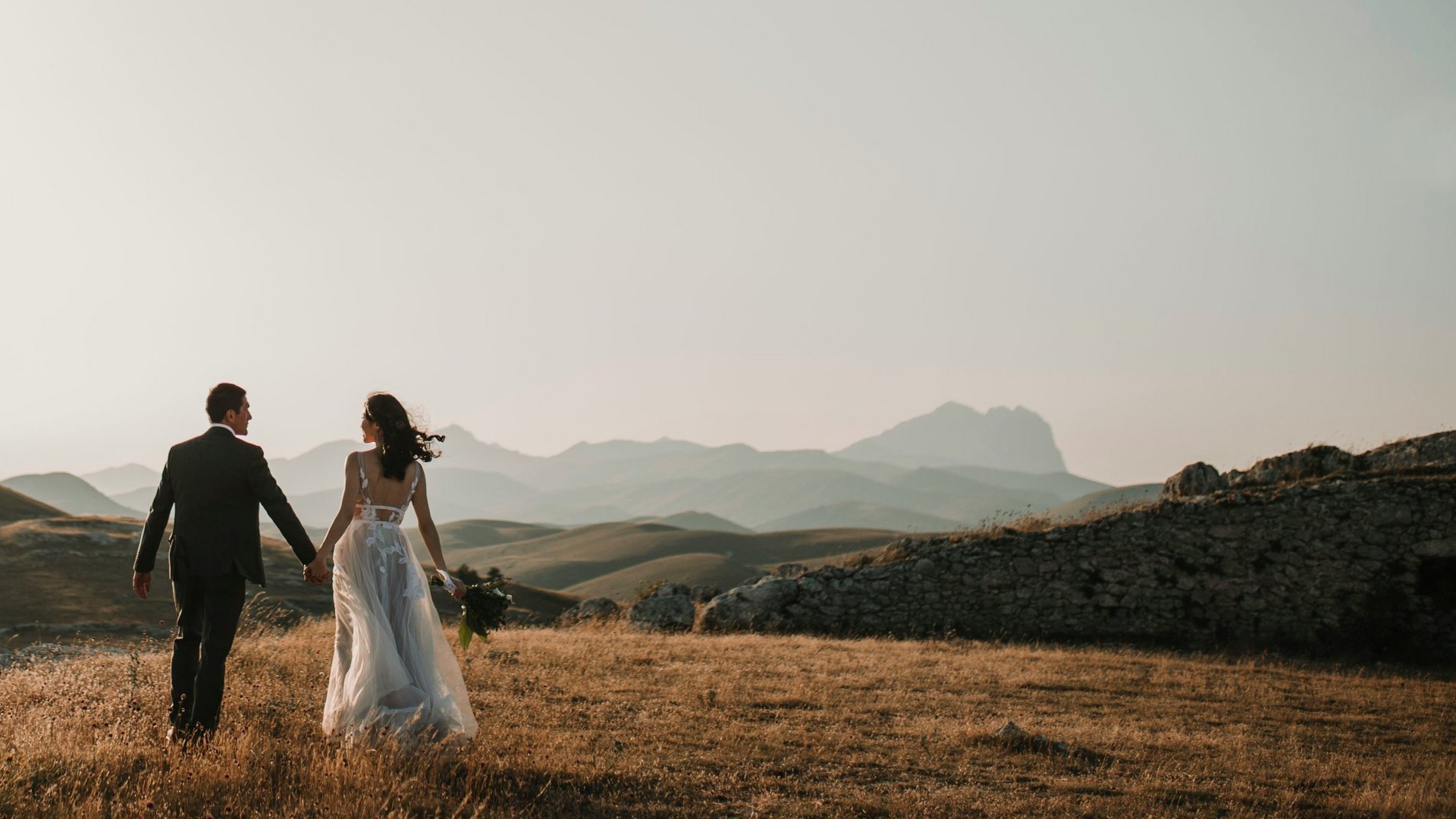 Paar in Hochzeitskleidung hält sich an der Hand auf einem Hügel bei Sonnenuntergang