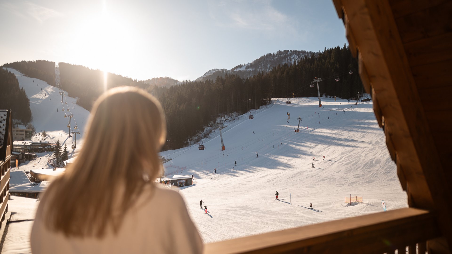Frau blickt von Holzbalkon auf sonnige Skipiste mit Skifahrern und Gondeln