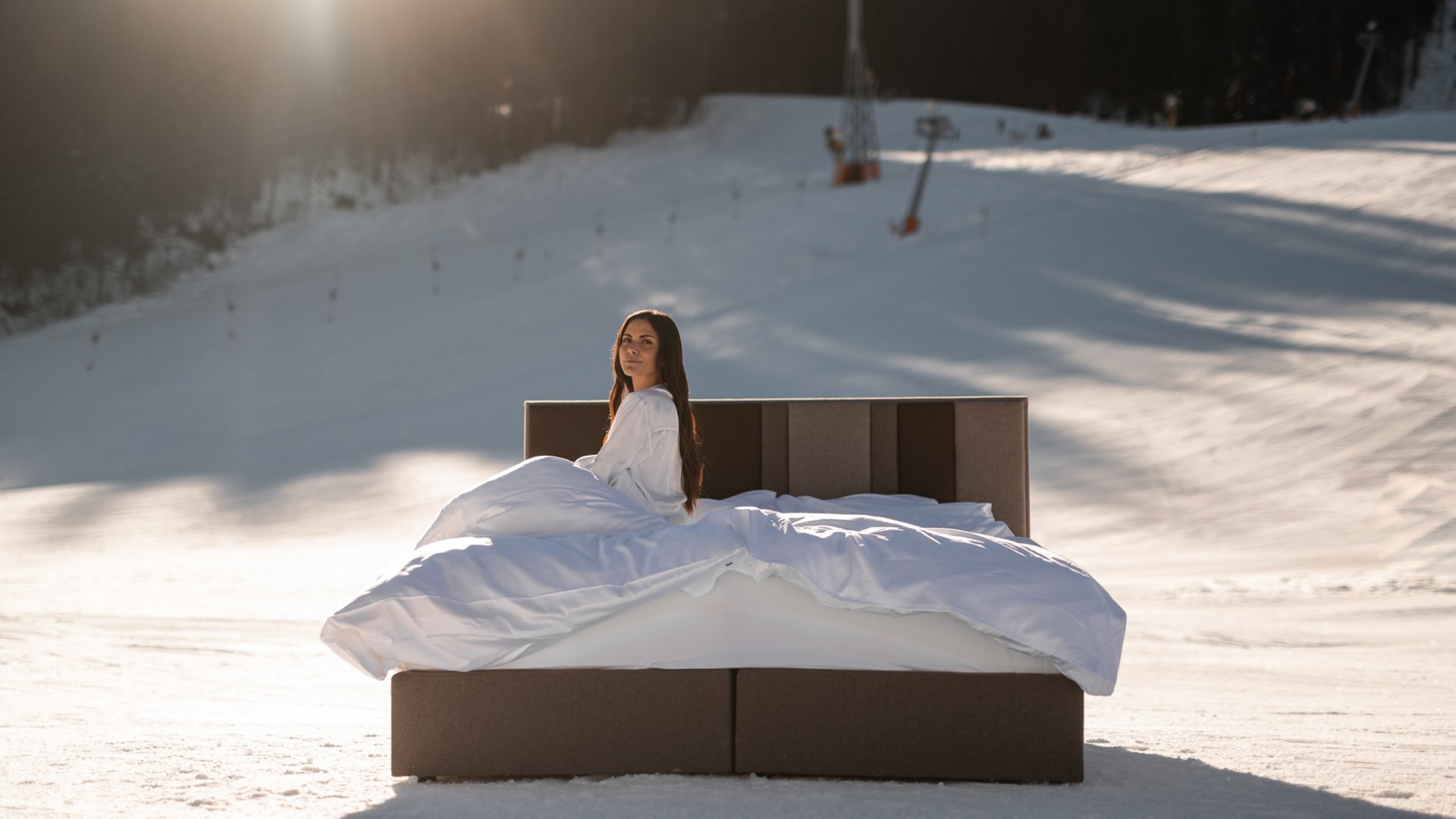 Frau sitzt in Bett mit weißen Decken auf verschneitem Hügel bei Sonnenlicht