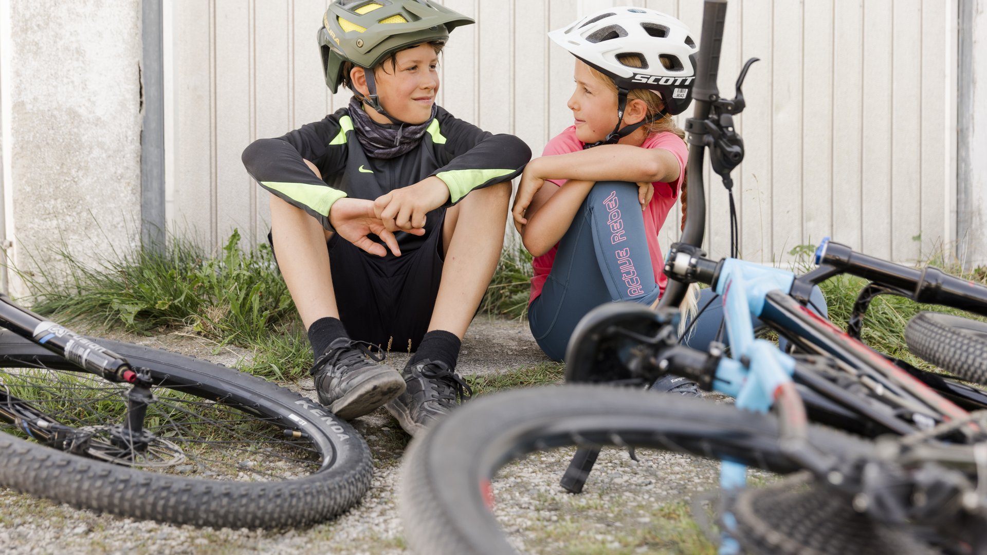 Zwei Kinder sitzen am Boden und lachen sich gegenseitig an, im Vordergrund sind ihre Mountainbikes Zwei Kinder sitzen am Boden und lachen sich gegenseitig an, im Vordergrund sind ihre Mountainbikes