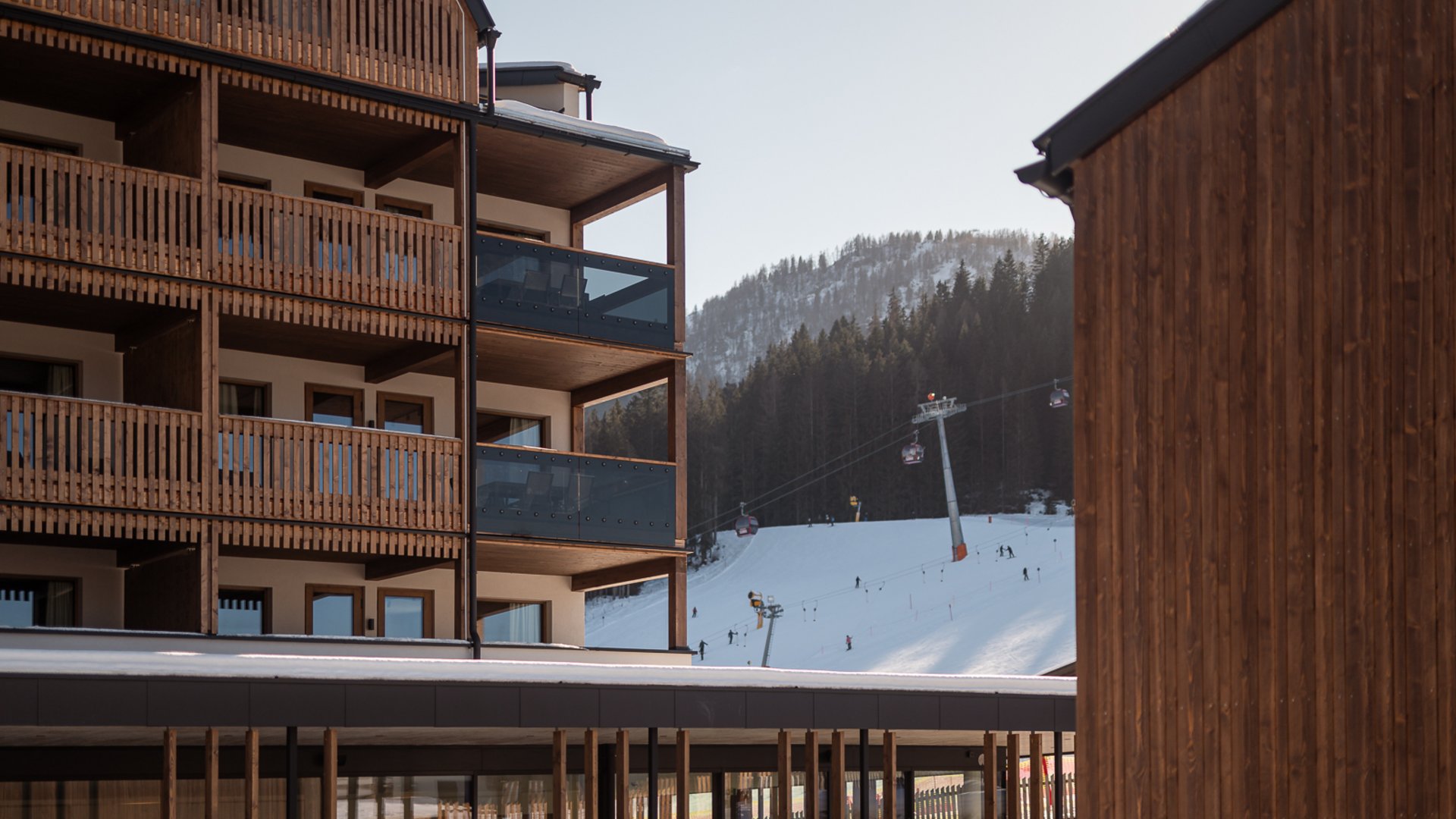 Hotel in Fieberbrunn: ADEA Lifestyle Suites Holzgebäude neben Skipiste mit Gondeln und schneebedecktem Berg