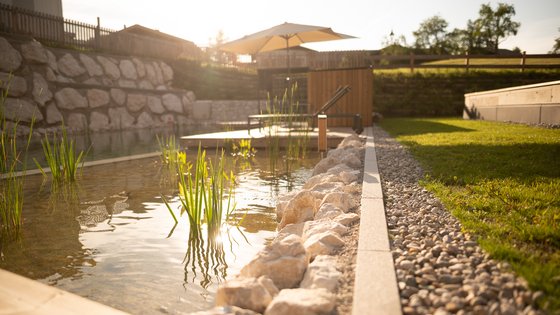 Hotel in Fieberbrunn: ADEA Lifestyle Suites Natürlicher Schwimmteich mit Steinen, Pflanzen und Sonnenschirm in sonnigem Garten