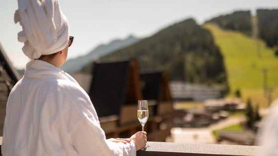 Frau steht im Bademantel und Handtuch in den Haaren auf dem Balkon mit einem Glas Sekt und schaut auf die Kitzbüheler Alpen Frau steht im Bademantel und Handtuch in den Haaren auf dem Balkon mit einem Glas Sekt und schaut auf die Kitzbüheler Alpen