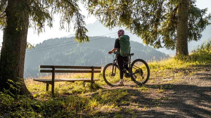 Alleinreisende Fahrradfahrerin mit Helm und Rucksack auf Waldweg neben Bank mit Bergblick