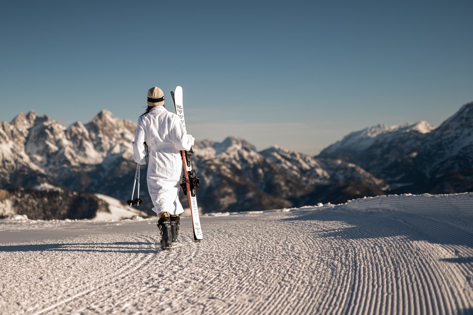 More mountains. More space. More time together. Person in weißem Skianzug trägt Ski und Stöcke auf schneebedeckter Piste mit Bergblick