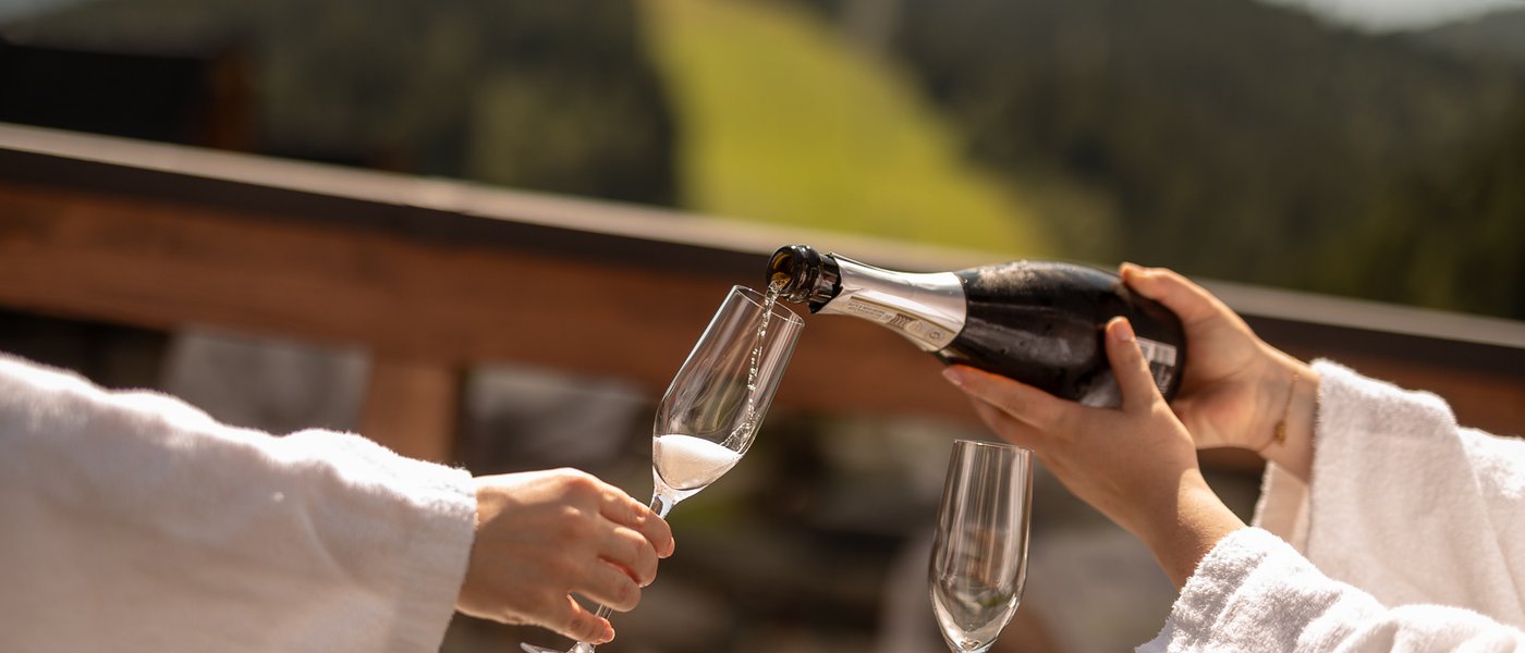 Mit Freundinnen in den Urlaub in Österreich Person gießt Sekt in Glas auf Terrasse mit Bergblick