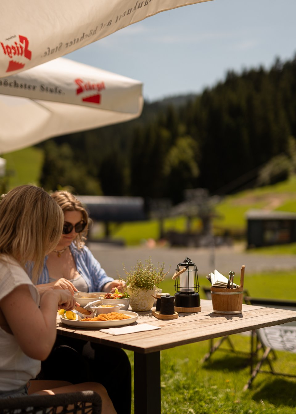 Hotel in Fieberbrunn: ADEA Lifestyle Suites Zwei Frauen essen im Freien an einem Holztisch unter Sonnenschirmen mit Bergkulisse