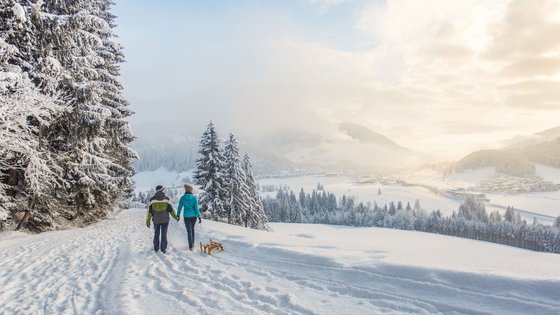 Kurzurlaub in Tirol: Wellness-Auszeit im ADEA Fieberbrunn Zwei Menschen gehen im Schnee mit einem Schlitten auf einem verschneiten Weg