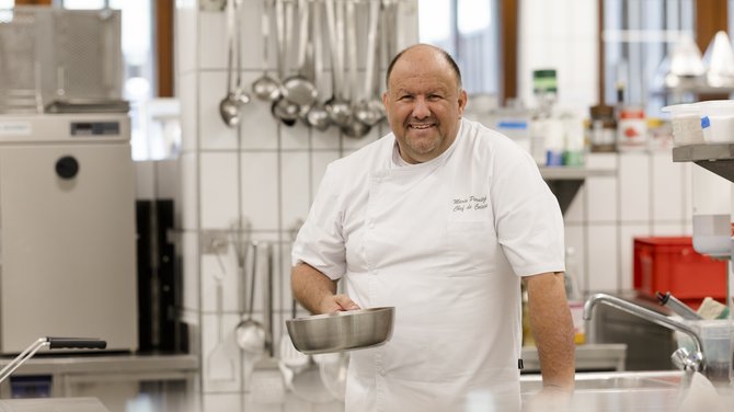 Team der ADEA Lifestyle Suites Fieberbrunn Lächelnder Koch in weißer Uniform hält Pfanne in professioneller Küche