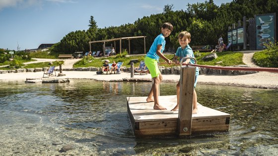 Kurzurlaub in Tirol: Wellness-Auszeit im ADEA Fieberbrunn Zwei Jungen ziehen eine Holzplattform mit Seil über klares Wasser am Strand