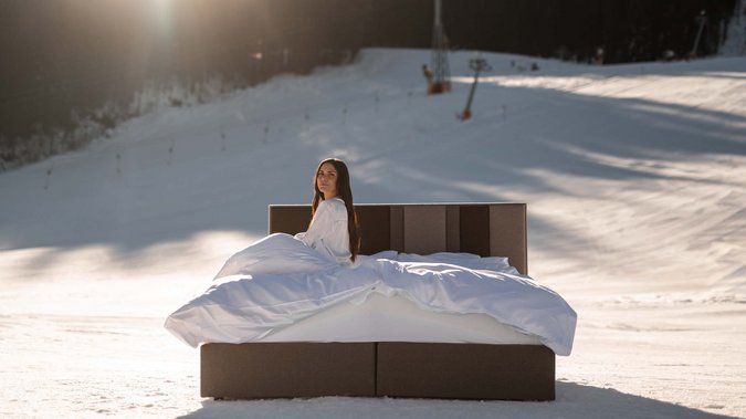 Hotel in Fieberbrunn: ADEA Lifestyle Suites Frau sitzt auf einem Bett im Schnee vor einem schneebedeckten Hang bei Sonnenlicht