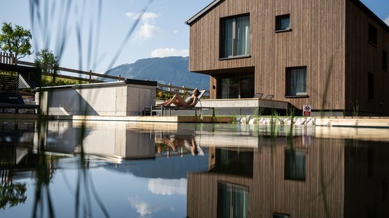 Hotel in Fieberbrunn: ADEA Lifestyle Suites Frau entspannt auf Liegestuhl neben modernem Holzhaus mit Teich und Bergblick