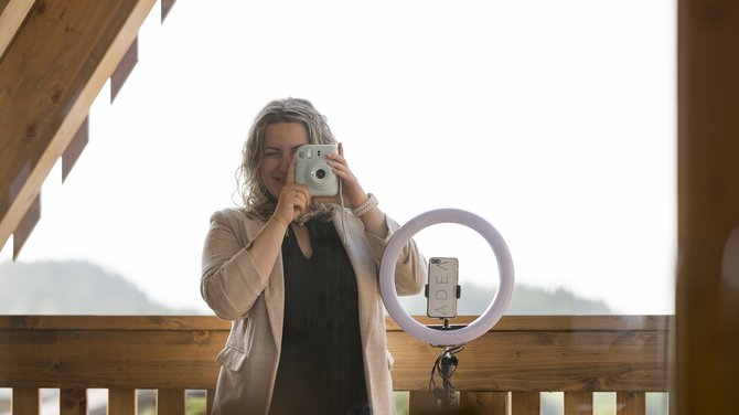 Team der ADEA Lifestyle Suites Fieberbrunn Frau nimmt Foto mit Sofortbildkamera auf einem Holzdeck mit Ringlicht und Smartphone