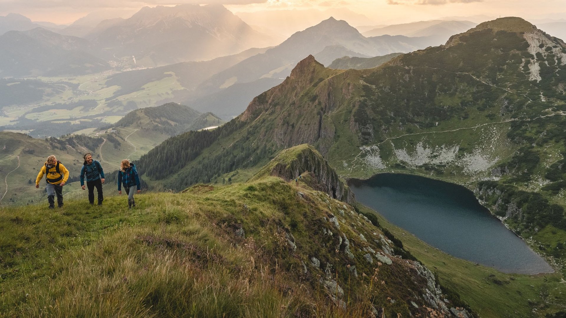 Kurzurlaub in Tirol: Wellness-Auszeit im ADEA Fieberbrunn Drei Wanderer steigen auf einem grasbewachsenen Berg mit See und Bergen im Hintergrund auf