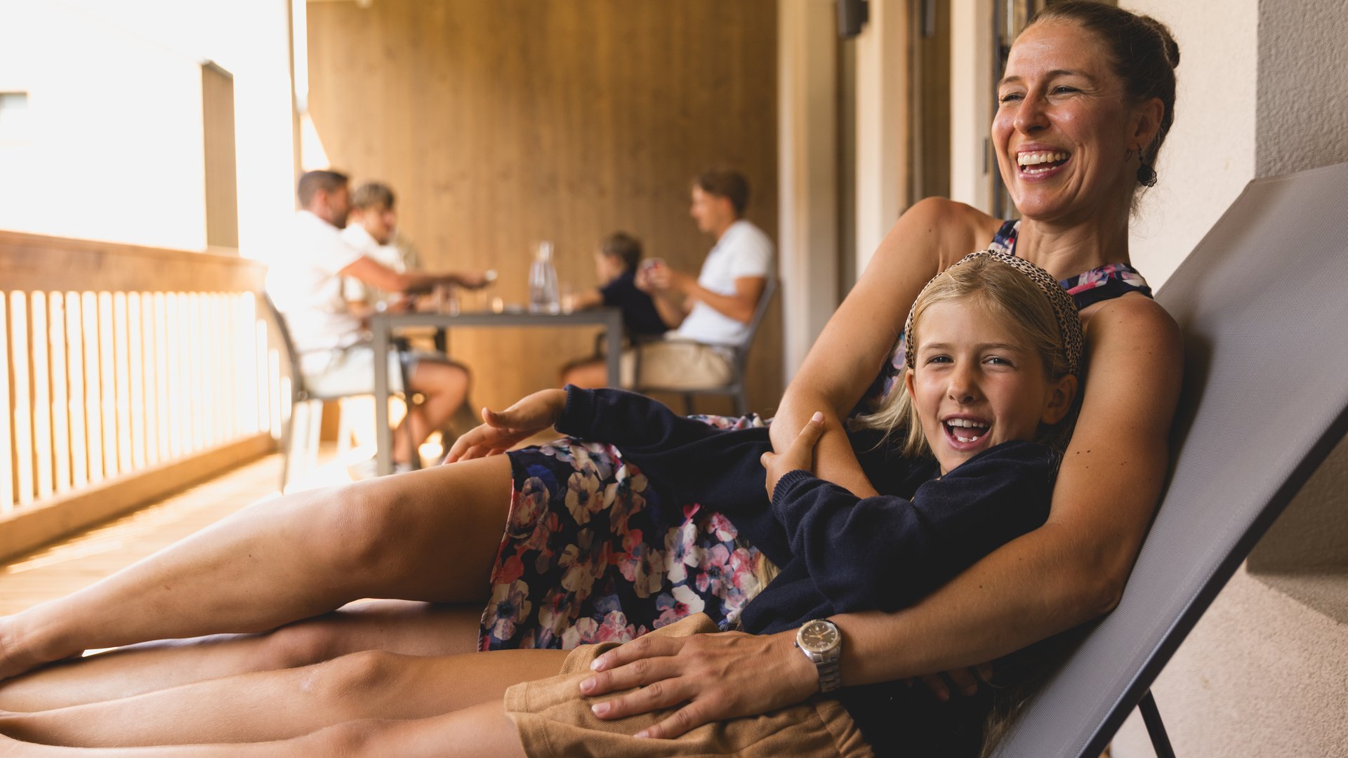 Mutter liegt mit Kind auf der Sonnenliege am Balkon, im Hintergrund sitzt die restliche Familie am Tisch Mutter liegt mit Kind auf der Sonnenliege am Balkon, im Hintergrund sitzt die restliche Familie am Tisch