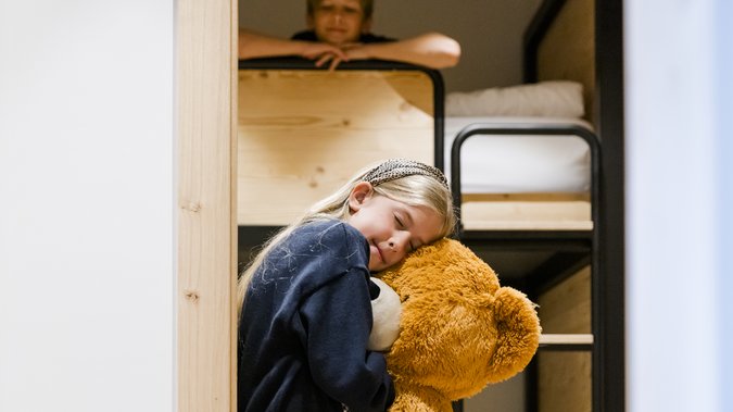 Girl holding a huge teddy bear, with a bunk bed in the background on which a boy is sitting. Girl holding a huge teddy bear, with a bunk bed in the background on which a boy is sitting.