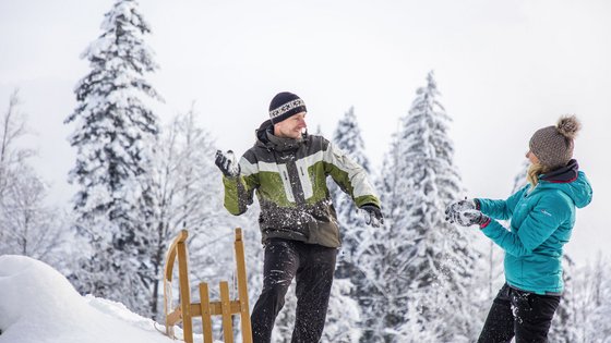 Kurzurlaub in Tirol: Wellness-Auszeit im ADEA Fieberbrunn Paar hat Schneeballschlacht im Schnee mit Schlitten und verschneiten Bäumen