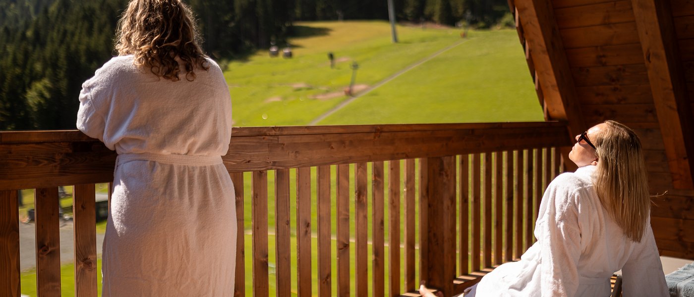 Mit Freundinnen in den Urlaub in Österreich Zwei Frauen in Bademänteln genießen die Aussicht auf grüne Berge von einem Holzbalkon