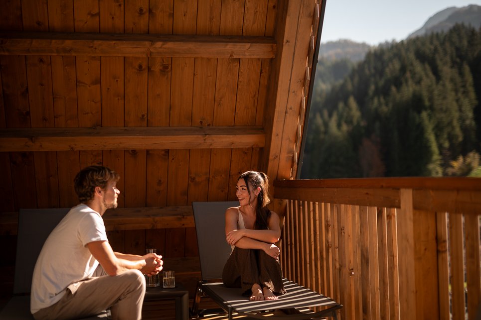 More mountains. More space. More time together. Paar entspannt auf Balkon einer Holzhütte mit Bergblick im Sonnenlicht