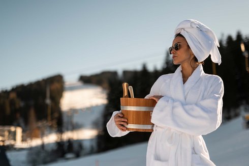 Woman standing on the ski slope wearing a bathrobe and holding a sauna infusion bucket in her hand Woman standing on the ski slope wearing a bathrobe and holding a sauna infusion bucket in her hand