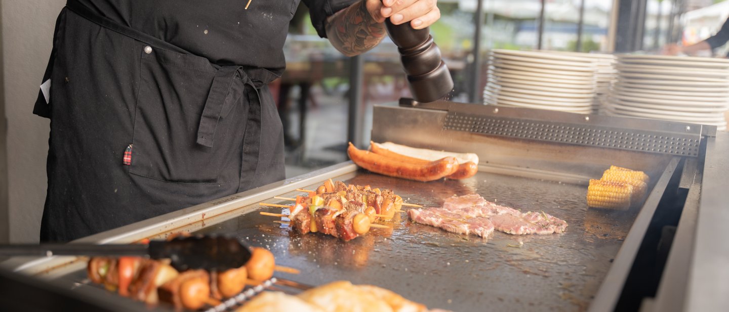Koch steht am Grill und würzt ein Fleisch, auf der Grillplatte liegen Würstchen, Fleisch und Hähnchenkeulen Koch steht am Grill und würzt ein Fleisch, auf der Grillplatte liegen Würstchen, Fleisch und Hähnchenkeulen